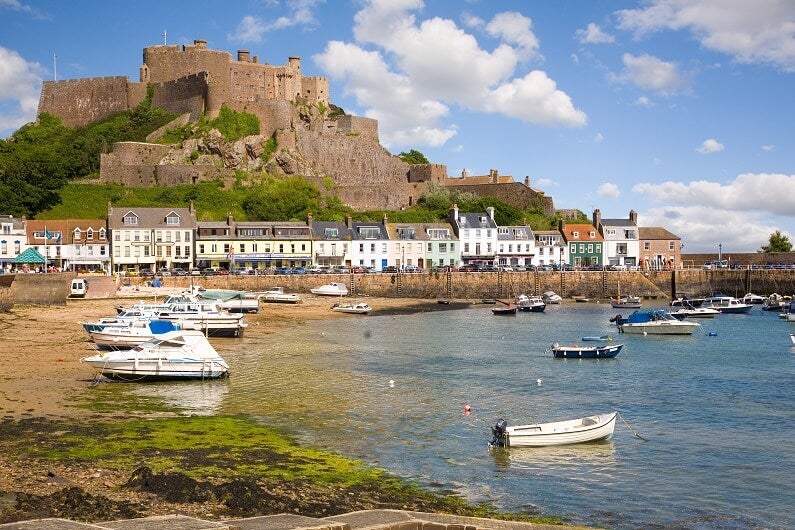 A coastal town in Jersey, with boats in the bay and a castle on a hill in the background.